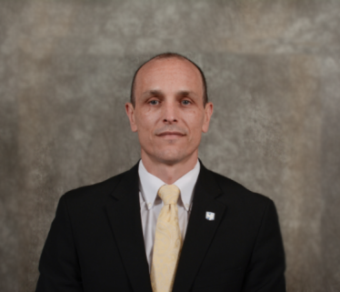 White bald man executive in black suit with yellow tie against a brown background