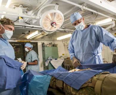 Two masked and gowned surgeons prepare for procedure on simulated service member.
