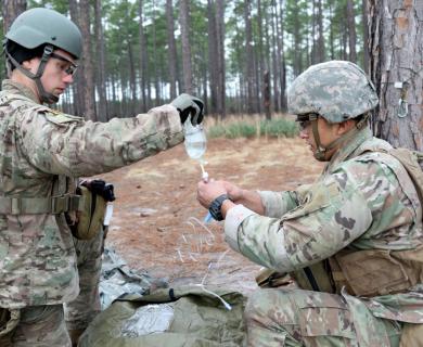 Two army medics work on a saline bag, outside in the forest. 