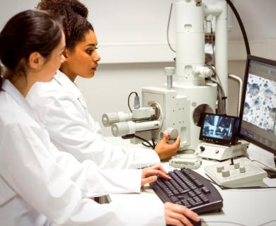 Two female scientists look at computer together while using microscope