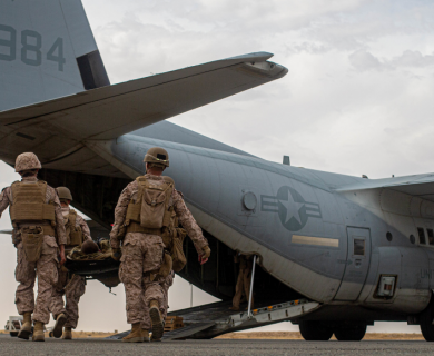 Four military personnel carry a stretcher toward a loading plane