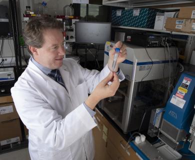 White man in lab coat holding beaker with lab in background