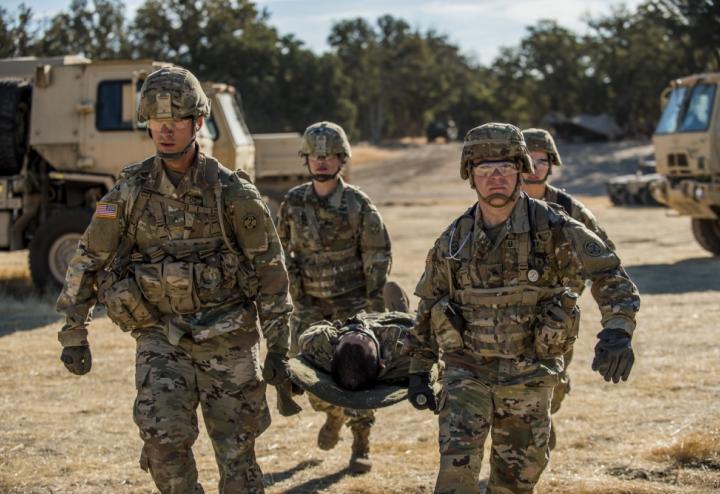 3 Army medics carry stretcher with simulated casualty. Vehicles are in the background.