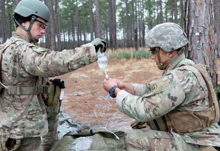 Two army medics work on a saline bag, outside in the forest.