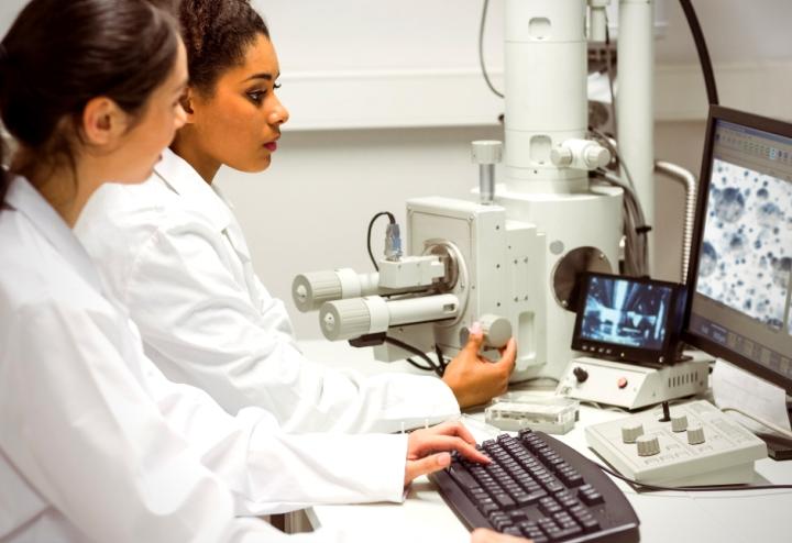 Two female scientists look at computer together while using microscope