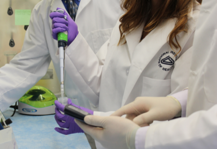 Two people in lab coats stand around a woman in a lab coat holding up a syringe. Source: National Institute of Health.