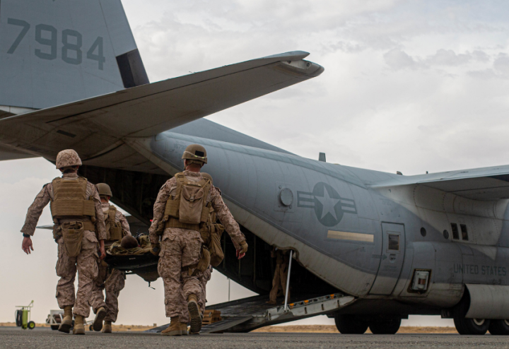 Four military personnel carry a stretcher toward a loading plane