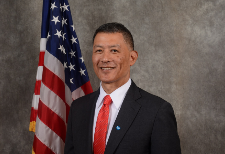 Asian American man in black suit with red tie, American flag in the background