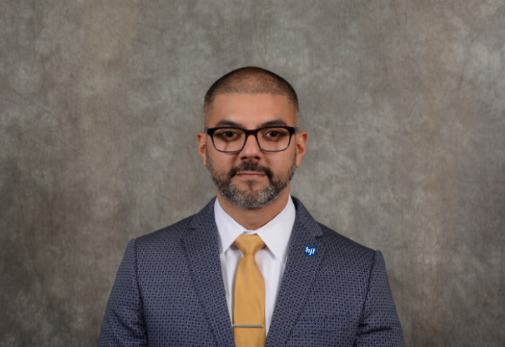 Man with glasses and beard in grey suit against brown background.