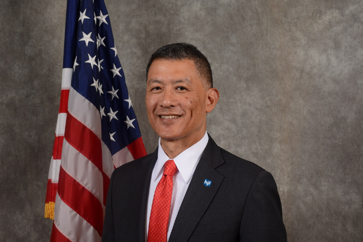 Asian American man in black suit with red tie, American flag in the background