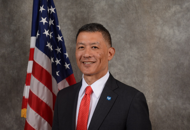 Asian American man in black suit with red tie, American flag in the background