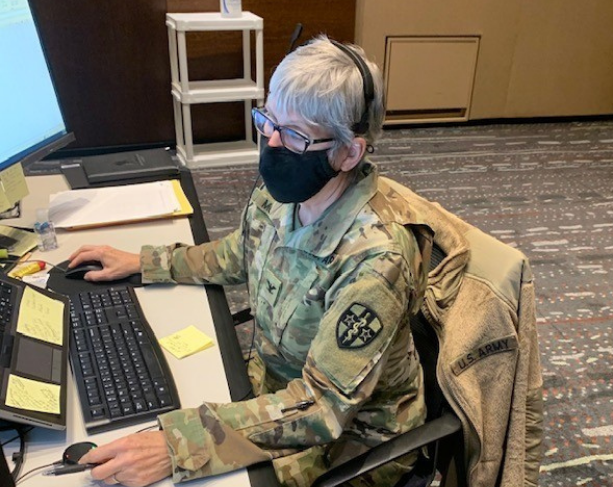 Older woman in military uniform and mask works on computer at her desk