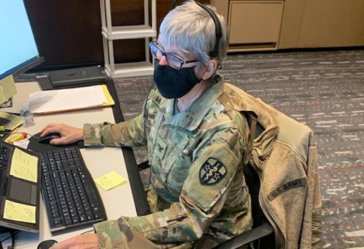 Older woman in military uniform and mask works on computer at her desk