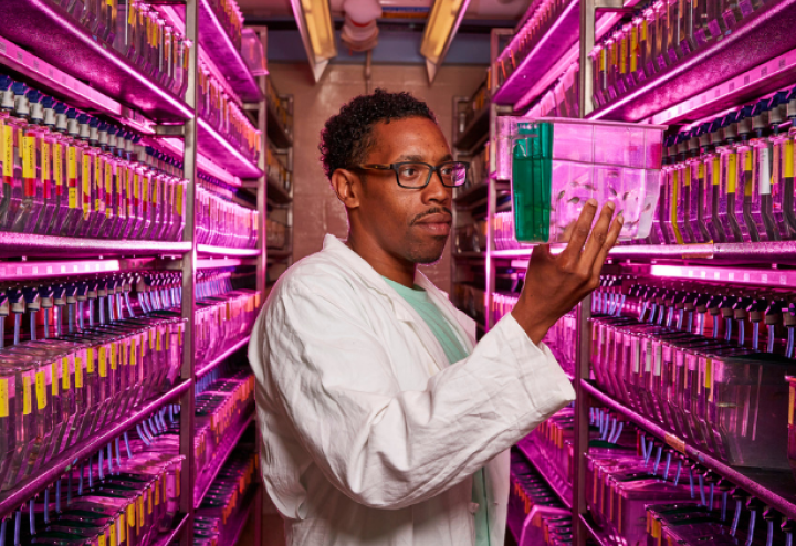 Black man in lab coat holds up research specimen in laboratory hallway of stored specimen. Source: NIH.