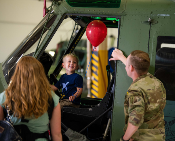Small blonde toddler sits in the driver's seat of a truck with a red balloon and laughs with his father in military uniform 