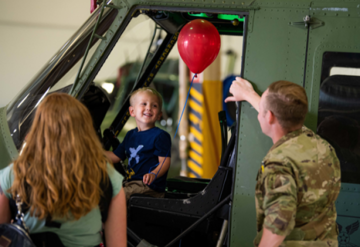 Small blonde toddler sits in the driver's seat of a truck with a red balloon and laughs with his father in military uniform 