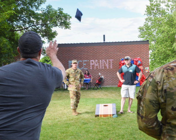 A man tosses a sack for a game of cornhole with a man in a miltary uniform on the other side and a Face Paint sign behind them
